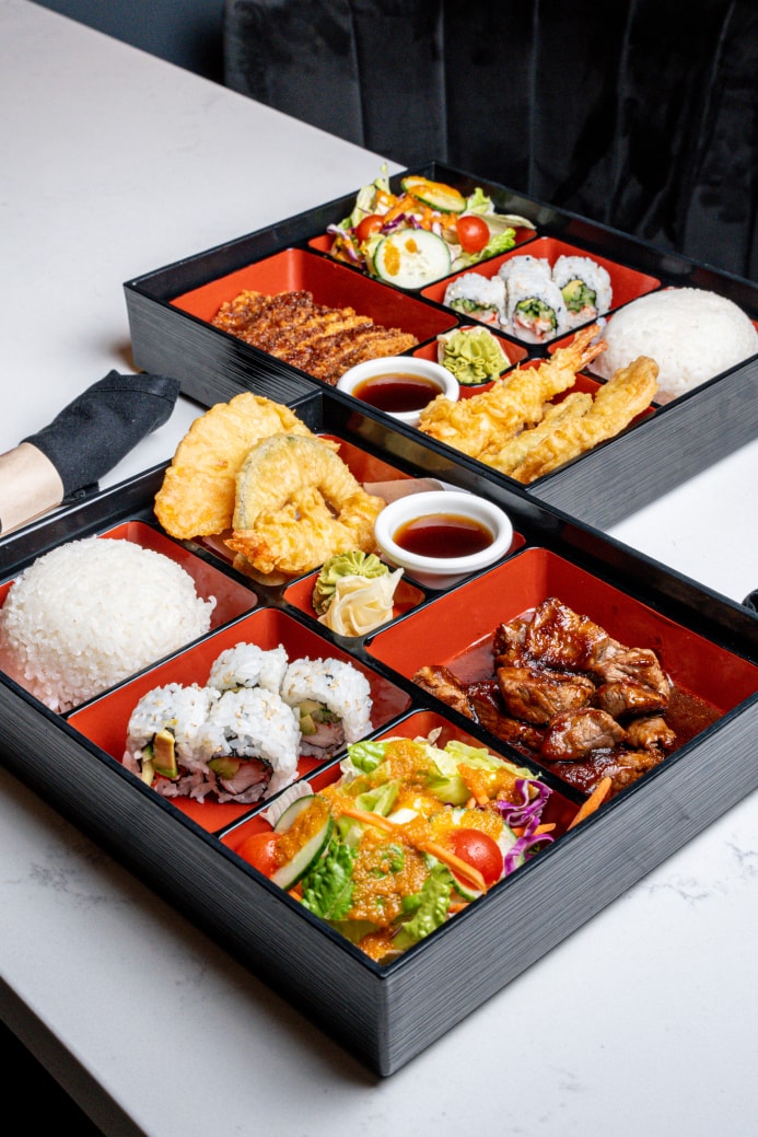 Two bento boxes with sushi rolls, shrimp tempura, teriyaki, rice and salad